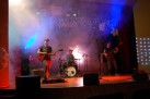 Zdjęcie przedstawia zespół muzyczny występujący na scenie. Na scenie znajduje się trzech muzyków: gitarzysta z gitarą elektryczną po lewej stronie, perkusista za zestawem perkusyjnym w centrum oraz basista z gitarą basową po prawej stronie. Scena jest oświetlona kolorowymi reflektorami, a w tle widać kurtynę i dekoracje ze światełek oraz balonów.