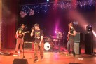 Zdjęcie przedstawia koncert zespołu muzycznego na scenie. Na scenie widać czterech muzyków: wokalistę, dwóch gitarzystów i perkusistę grającego na perkusji. Scena jest oświetlona kolorowymi światłami i ozdobiona girlandami światełek.
