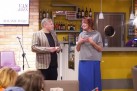 Zdjęcie przedstawia mężczyznę zwracającego się w stronę kobiety.  Kobieta zaś splata dłonie na piersiach. Za nimi bar. Widać także kilka osób z publiki, wyświetlacz na ceglastej ścianie, krzesło i mikrofon. 