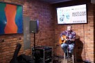 Na zdjęciu mężczyzna. Siedzi na wysokim krześle, gra na gitarze i śpiewa do mikrofonu. Za nim na ekranie wyświetlana jest grafika dotycząca odbywającego się wydarzenia. Widać również sprzęt muzyczny ustawiony przed gitarzystą oraz duży obraz wiszący na ścianie. 