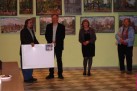 Na zdjęciu dwie kobiety i dwaj mężczyźni na tle obrazów wiszących na ścianie. Jeden z mężczyzn trzyma w dłoni duży, biały artykuł w kształcie prostokąta. 