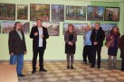 Zdjęcie prezentuje kilka osób, zarówno kobiety jak i mężczyźni stojący przed ścianą, na której wiszą obrazy. 