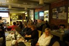  Publiczność siedząca przy stolikach podczas koncertu. W tle muzyk dający koncert.