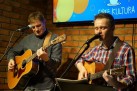 Na zdjęciu dwaj mężczyźni grający na gitarach. Jeden prawdopodobnie śpiewa do mikrofonu. 