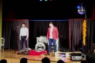 Zdjęcie przedstawia występ kabaretu Hrabi. Na scenie widać trzech mężczyzn jeden z mężczyzn leży na podłodze. Widać także część publiczności. Na bębnie perkusyjnym umieszczone jest logo grupy. 