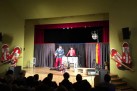 Na zdjęciu widać przedstawienie lub spektakl na scenie, z publicznością siedzącą na widowni. Na scenie znajdują się dwie osoby w kostiumach oraz trzecia osoba przy konsoli DJ-skiej. Scena jest oświetlona i udekorowana czerwonymi zasłonami oraz dużymi, stylizowanymi figurami na ścianach po bokach.