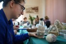 Na pierwszym planie osoba spoglądająca na ozdobione jajo wielkanocne. W tle widać innych uczestników wydarzenia oraz przedmioty znajdujące się na stołach. 