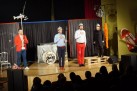 Na zdjęciu widać występ kabaretowy. Czterech artystów stoi na scenie przed publicznością. Na scenie znajduje się bęben z napisem „hrabi”, keyboard, krzesło i dekoracje. Artyści występujący trzech mężczyzn i jedna kobieta. Wszyscy mają na głowach hełmy, a ich stroje wyglądają na stylizowane. Jeden z mężczyzn trzyma długi kij z rybą. W dolnej części zdjęcia widać publiczność siedzącą w rzędach, oglądającą występ. 