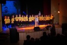 Na zdjęciu grupa dzieci ubranych w ciemne spodnie i żółte koszulki, stoją w rzędzie. Jedna osoba przebrana jest za zwierzątko i stoi przed resztą dzieci. Widać część publiczności. Światło oświetlające scenę ma kolor żółty i niebieski..  