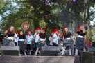 Zdjęcie przedstawia grupę dzieci występujących na scenie. Dzieci tańczą na scenie plenerowej, za którą widać drzewa. Na scenie znajdują się głośniki i inny sprzęt nagłośnieniowy. Dzieci ubrane są w stroje taneczne. Stroje dziewczynek składają się z czarnych bluzek, czarno-białych spódnic w kropki oraz czerwonych czapek z kwiatami, zaś chłopcy mają założone białe bluzki z czerwonymi muszkami, czarne spodnie, czerwone kapelusze, a na dłoniach białe rękawiczki. 
