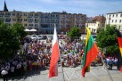 Na zdjęciu widać duży tłum ludzi zgromadzonych na placu. W centralnej części kadru znajduje się duża grupa ludzi, w tym dzieci i dorośli. Niektórzy mają na sobie kolorowe stroje lub trzymają transparenty. Na pierwszym planie widoczne są flagi, w tym flaga Polski (biało-czerwona) oraz flaga Litwy (żółto-zielono-czerwona). Po prawej stronie widać również fragment flagi Niemiec (czarno-czerwono-złota). W tle znajdują się budynki mieszkalne oraz inne obiekty. Na placu widać również zaparkowane samochody. Zdjęcie zostało zrobione w słoneczny dzień.