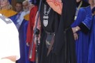Na zdjęciu widać mężczyznę w stroju ceremonialnym, który przemawia do mikrofonu. Mężczyzna w średnim wieku, w czarnej todze z elementami w kolorze ceglastym i czarnym, oraz w czarnym birecie. Na szyi ma łańcuch, a przy pasie sakiewkę i sztylet. Mówi do mikrofonu na statywie. Za mężczyzną stoi grupa osób, również ubranych w togi. Kobieta po prawej stronie ma niebieską togę i okulary. Tło stanowią budynek z oknami oraz fragmenty innych osób.
