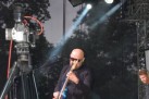 Na zdjęciu łysy mężczyzna w czarnym stroju i okularach przeciwsłonecznych gra na niebieskiej gitarze elektrycznej. Scena jest oświetlona reflektorami, z tyłu widoczne są drzewa. Obok muzyka znajduje się kamera na statywie, a po prawej stronie widoczny jest fragment klawiatury marki Yamaha. Po prawej stronie widać też drugą osobę, prawdopodobnie innego muzyka.