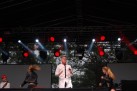 Zdjęcie przedstawia występ muzyczny na scenie. W centrum znajduje się wokalista w białej koszuli, a po jego bokach dwie tancerki w czarnych strojach. Scena jest oświetlona reflektorami w kolorach czerwonym i białym, a w tle widać resztę sceny oraz drzewa.