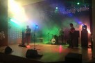Na zdjęciu widać zespół muzyczny występujący na scenie. Scena jest oświetlona kolorowymi reflektorami. W powietrzu unosi się dym. W tle widać dekoracje świetlne. Na scenie znajdują się trzej muzycy. Jeden z nich gra na gitarze elektrycznej po lewej stronie, drugi na perkusji w centralnej części, a trzeci, również z gitarą, stoi po prawej stronie. Na scenie widoczne są głośniki, wzmacniacze gitarowe, statywy mikrofonowe oraz zestaw perkusyjny.