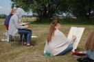 Obraz przedstawia grupę osób siedzących na trawie na świeżym powietrzu, prawdopodobnie podczas pleneru malarskiego lub zajęć z rysunku. Uczestnicy siedzą na krzesłach lub bezpośrednio na ziemi i szkicują na kartkach papieru umieszczonych na podkładkach. W tle widać drzewa, pole i domy.