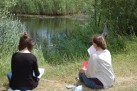 Zdjęcie przedstawia dwie osoby siedzące na trawie, tyłem do obiektywu, nad brzegiem zbiornika wodnego. Obie osoby trzymają w rękach kartki lub szkicowniki. Wokół zbiornika rośnie bujna roślinność, w tym trzciny. 