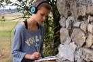 Zdjęcie przedstawia młodą kobietę, która siedzi na kamiennym murze na świeżym powietrzu. Słucha muzyki przez słuchawki, a jednocześnie pisze lub rysuje w otwartym zeszycie. W tle widać drzewo i otwartą przestrzeń.