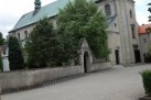 Zdjęcie przedstawia kościół. Widać na nim fasadę świątyni z wysoką wieżą i zielonym dachem. Przed kościołem, na brukowanym placu, siedzą dwie osoby, które rysują lub malują.