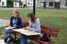 Zdjęcie przedstawia dwie młode dziewczyny siedzące na ławce w parku lub na placu. Obie są pochylone nad swoimi notatnikami lub blokami do rysowania. W tle widać budynki mieszkalne, a w oddali inne osoby.