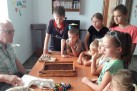 Na zdjęciu dzieci zgromadzone wokół stołu, przy którym siedzi starszy mężczyzna. Na stole znajduje się kilka przedmiotów, m.in. książka.