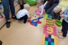 Na zdjęciu znajduje się grupa małych dzieci bawiących się na podłodze oraz jedną dorosłą osobę (prawdopodobnie opiekunkę lub rodzica), która kuca obok dzieci. Dzieci bawią się małymi figurkami i zabawkami. Na podłodze leżą również kolorowe, piankowe maty z cyframi. Akcja rozgrywa się w pomieszczeniu z jasną podłogą i regałami z książkami w tle.