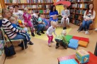 Osoby dorosłe i dzieci w bibliotece. Kobiety siedzą na krzesłach i trzymają na kolanach dzieci. Dwójka dzieci bawi się dużymi kolorowymi klockami. W tle widać regały z książkami.