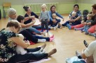Na zdjęciu widać grupę kobiet, siedzących na podłodze w pomieszczeniu i zajmujących się małymi dziećmi. Dorośli siedzą w kręgu na macie lub kocach rozłożonych na drewnianej podłodze i wchodzą w interakcje z dziećmi.