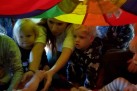 Na zdjęciu widać grupę małych dzieci i dorosłych bawiących się pod kolorową chustą animacyjną.