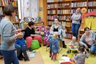 Fotografia prezentuje spotkanie edukacyjne lub warsztaty dla dzieci i ich rodziców/opiekunów zorganizowane w bibliotece. W wydarzeniu uczestniczy grupa dorosłych i małych dzieci, którzy wspólnie spędzają czas w specjalnie zaaranżowanym kąciku czytelniczym, otoczeni regałami z książkami.