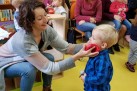 Zdjęcie przedstawia kobietę klęczącą przed małym chłopcem z blond włosami, który ma na sobie niebieską koszulę w kratę. Kobieta trzyma czerwony przedmiot przy jego twarzy, a oboje uśmiechają się. W tle widać inne dzieci i dorosłych, siedzących na krzesłach, a także regały z książkami.