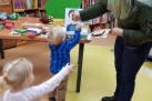 Zdjęcie przedstawia dorosłą osobę i dwoje małych dzieci w pomieszczeniu przypominającym bibliotekę lub salę zabaw. Dorosła osoba i jedno z dzieci stoją przy stole i układają puzzle lub elementy na dużej planszy z wizerunkiem twarzy. Drugie dziecko, dziewczynka, stoi z boku i wyciąga rękę w kierunku stołu. Na stole i w tle widać regały z książkami oraz inne zabawki i materiały edukacyjne.