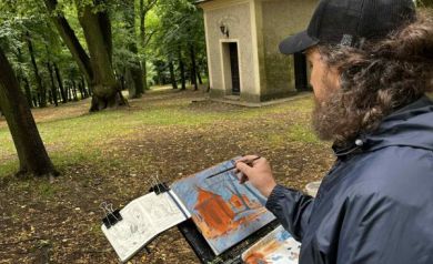 Mężczyzna malujący obraz. Na specjalnym stojaku znajduje się obraz, farby oraz inne przyrządy potrzebne do tworzenia dzieła. Mężczyzna na głowie ma czapkę z daszkiem, ubrany jest w kurtkę, w ręce trzyma pędzel, którym maluje. W tle widać drzewa i kaplicę.
