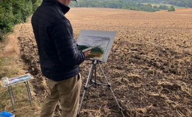 Mężczyzna ubrany w czapkę z daszkiem, ciemną kurtkę i jasne spodnie maluje widok, który się przed nim rozpościera. Obok niego różne sprzęty potrzebne do malowania. W tle widać pola i lasy. Niebo jest pokryte chmurami.