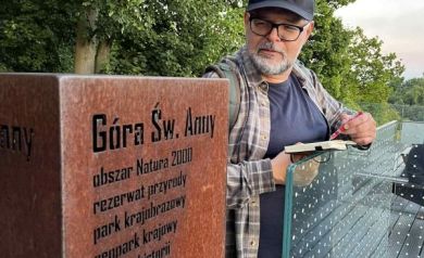 Mężczyzna w czarnych okularach, ubrany w czapkę z daszkiem, ciemną bluzkę i spodnie, a także w koszulę w kratkę stoi na tarasie widokowym i opiera się o szklaną barierkę. W rękach trzyma notes i długopis. Wpatruje się na tablicę, na której wypisane są skarby przyrody i dziedzictwa Góry Świętej Anny.