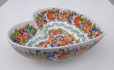 Na zdjęciu znajduje się ceramiczna miska w kształcie serca. Miska jest biała i ozdobiona kolorowym, tradycyjnym, kwiatowym wzorem w stylu ludowym, z przewagą czerwonych, niebieskich, pomarańczowych i zielonych motywów roślinnych. Miska stoi na jasnym, neutralnym tle. - powiększ