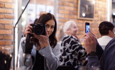 Kobieta pozuje do zdjęcia z aparatem w ręce na tle fotografii wiszących na ścianie.