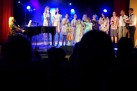 Na zdjęciu grupa śpiewających osób do mikrofonów. Jedna z kobiet gra na fortepianie, zaś jeden z mężczyzn na gitarze. Widać część przyciemnionej publiczności oraz niebieskie światła oświetlające scenę. 
