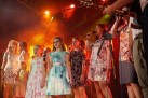 Zdjęcie osób śpiewających do mikrofonów. Jedna z osób gra na gitarze. Scenę oświetla czerwone i pomarańczowe światło. 