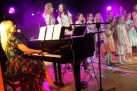 Na zdjęciu grupa śpiewających osób do mikrofonów i kobieta grająca na fortepianie. Scenę oświetla fioletowe światło. 