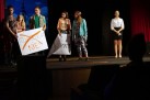 Na scenie aktorzy podczas odgrywania sceny. W dłoniach trzymają duże białe kartki. Na jednym przekreślony jest na pomarańczowo napis ,,TECHNOLOGIA' poniżej pomarańczowym kolorem napisane jest słowo ,,NIE!'. Obok grupy aktorów stoi młoda kobieta w stroju galowym. 