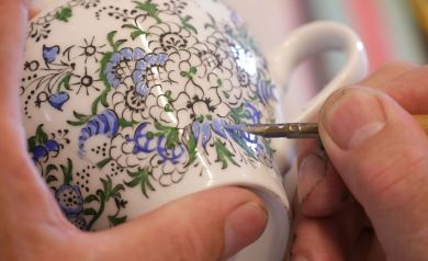 Na zdjęciu widoczna jest ręka osoby malującej precyzyjnie wzór kwiatowy na białym ceramicznym kubku. Wzór składa się z czarnych konturów i wypełniony jest niebieskimi oraz zielonymi detalami. Osoba używa cienkiego pędzelka do nanoszenia farby na powierzchnię kubka. Zdjęcie pokazuje proces ręcznego zdobienia ceramiki. - powiększ