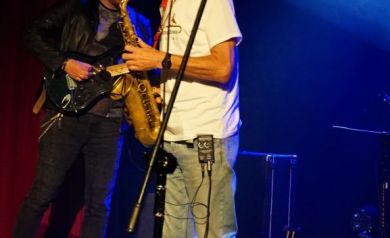Na scenie mężczyzna gra na saksofonie. Towarzyszy mu grający na gitarze muzyk. W tle widoczne jest światło o odcieniu niebieskim.