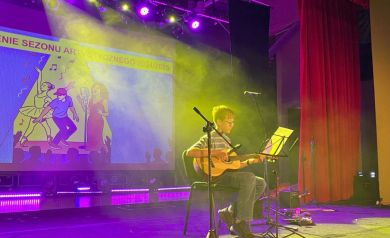 Na scenie chlopiec grający na gitarze. Wokół niego sprzęt muzyczny: mikrofony, stojak na nuty. W tle kolorowe światła i wyświetlany plakat odbywającej się imprezy. Widać również dużą czerwoną kotarę. 