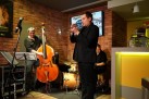Na zdjęciu trzech mężczyzn grających na instrumentach (trąbka, kontrabas i perkusja). Znajdują się w restauracji, gdzie ściany przypominają cegłę. Na jednej z ścian znajduje się ekran z wyświetlanym plakatem odbywającego się wydarzenia. 