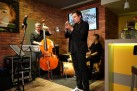Na zdjęciu trzech mężczyzn grających na instrumentach (trąbka, kontrabas i perkusja). Znajdują się w restauracji, gdzie ściany przypominają cegłę. Na jednej z ścian znajduje się ekran z wyświetlanym plakatem odbywającego się wydarzenia. Widać również część baru.