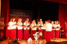 Na zdjęciu zespół śpiewaczy VIVA LA MUSICA podczas swojego występu. Śpiewający ubrani są w białe koszule i czerwone spódnice. Jedna kobieta dyryguje zespołem. Za występującymi wyświetlany jest napis ,,20-LECIE ZESPOŁU VIVA LA M,USICA'. 