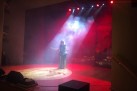 Na scenie kobieta ubrana w elegancką suknię. Występuje na scenie. Scena oświetlona jest czerwonymi światłami, unosi się tam również ciężki dym. 