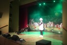 Na scenie dwóch aktorów w przebraniach. Za nimi scenografia przedstawiająca drzewa i polanę z kwiatami. Scenę oświetla zielone światło. 