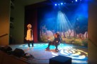 Na scenie dwóch aktorów w przebraniach. Za nimi scenografia przedstawiająca drzewa i polanę z kwiatami. Scenę oświetla niebieskie światło. 